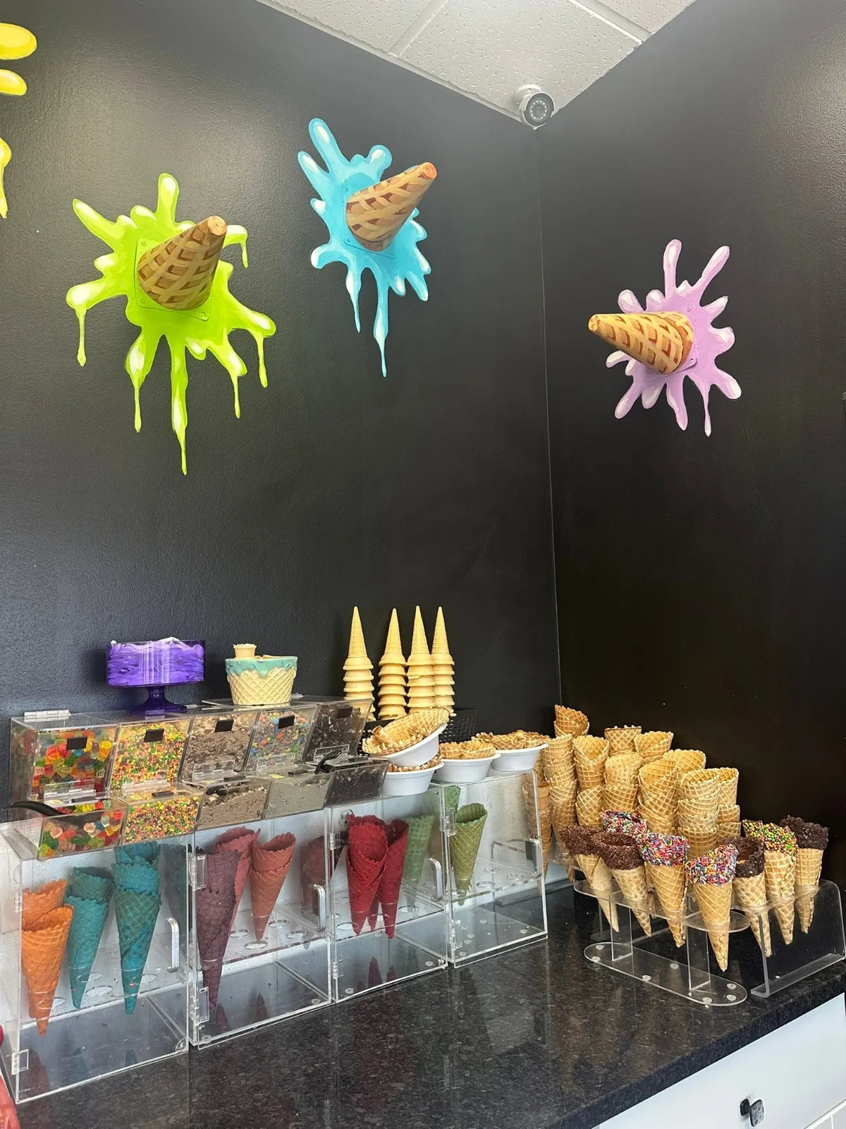 Waffle cones in rows on the counter, some dipped in chocolate and sprinkles, under a black wall painted with bright splashes
